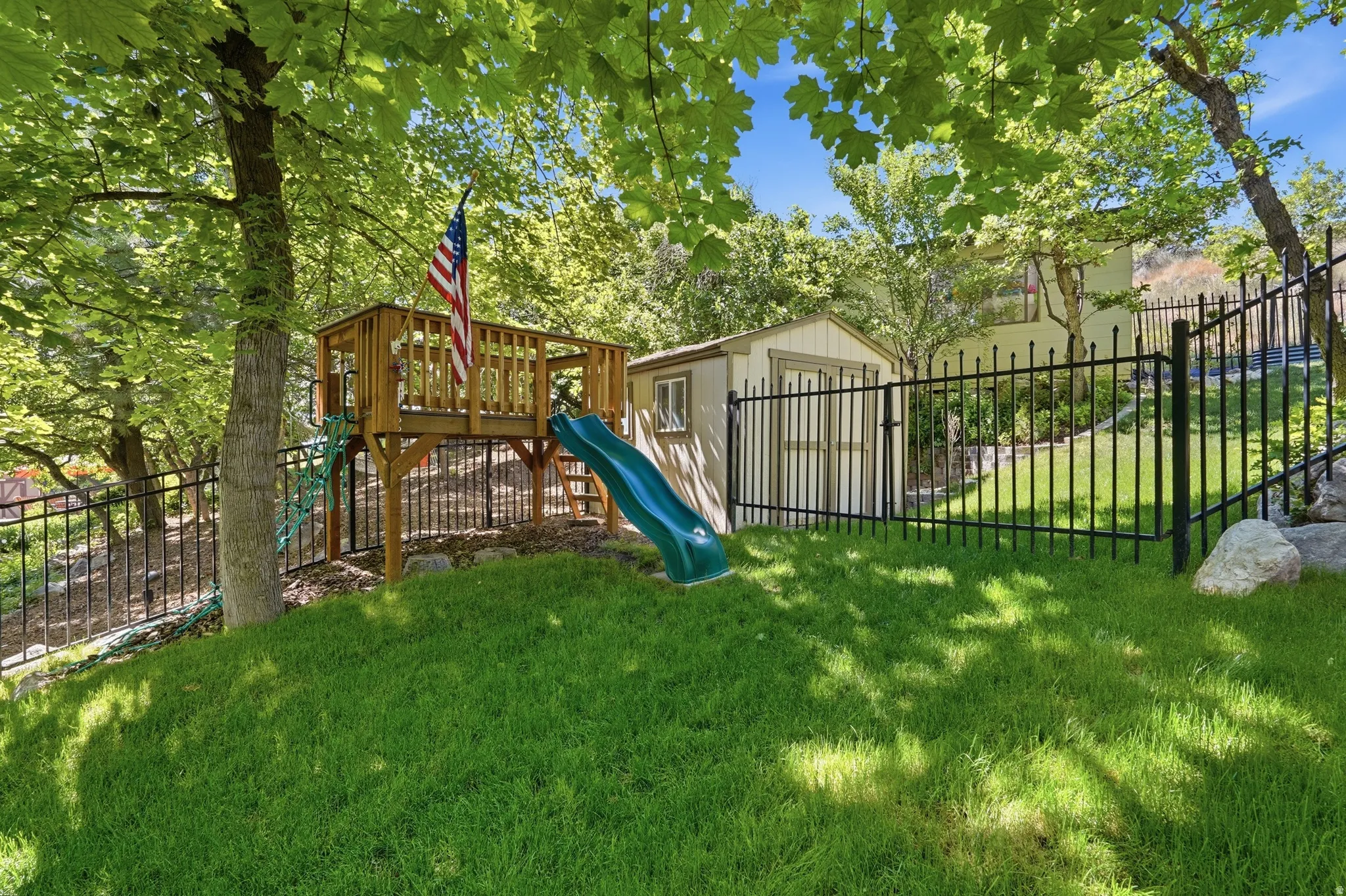 Sloped lawn area featuring a wooden play structure with a slide, a detached storage shed, and black metal fencing