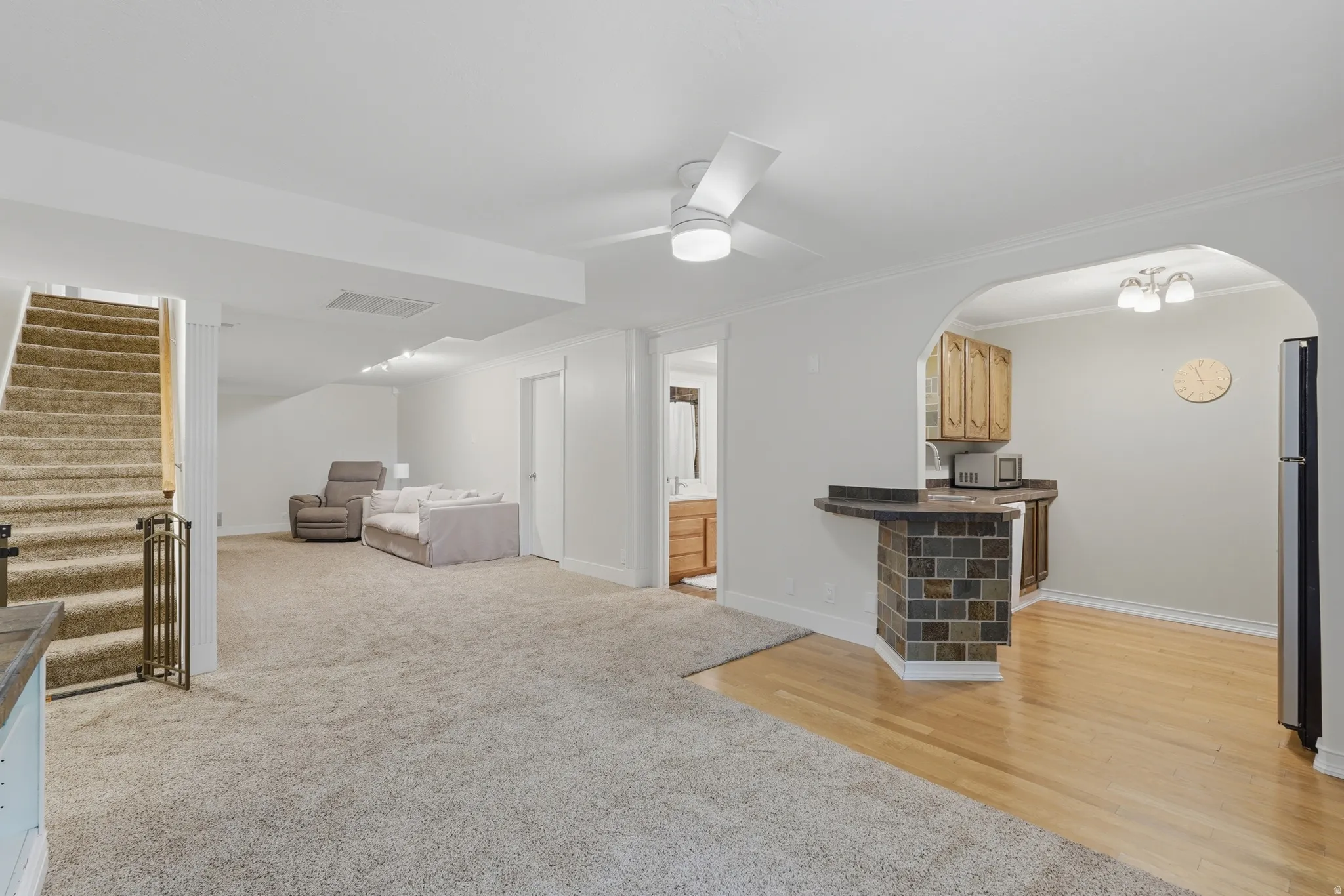 Open-concept living space with light-toned carpet flooring and a corner wet bar featuring a stacked stone base and dark countertop