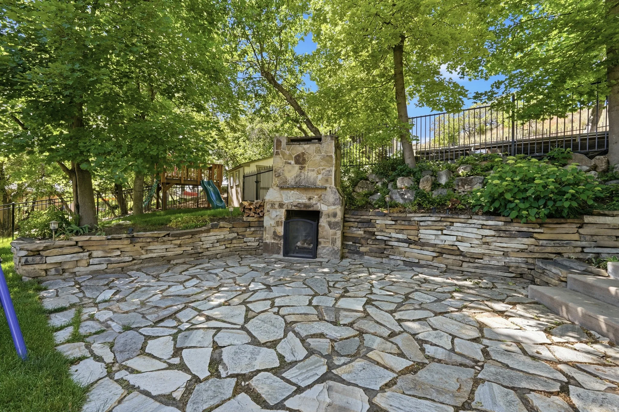 Stone flagstone patio with a built-in stone fireplace, featuring stacked stone retaining walls, mature trees, and a black wrought iron fence