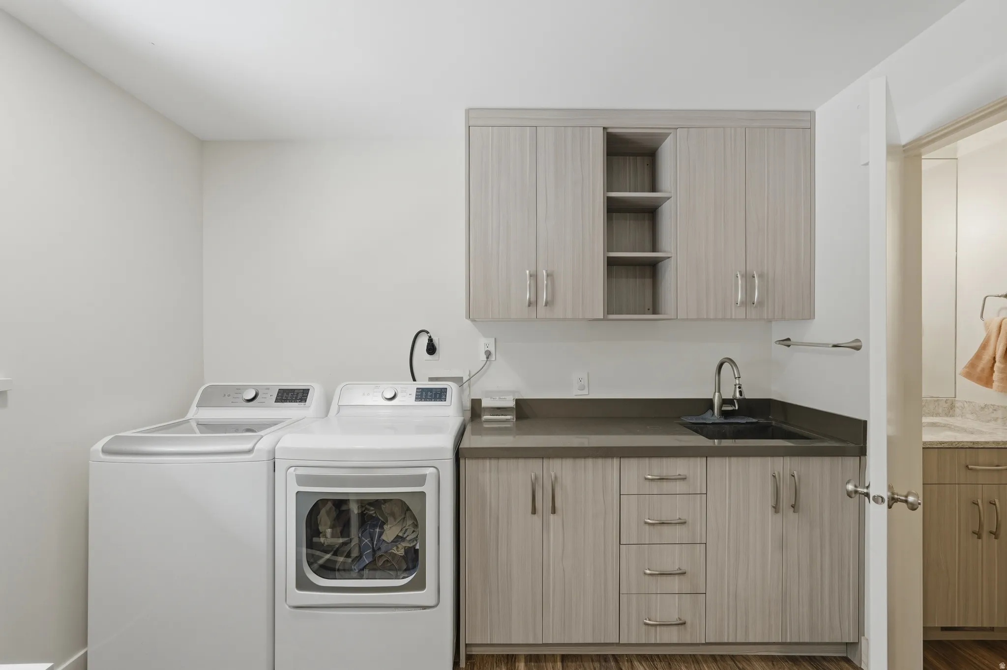 Dedicated laundry area featuring wood-finish flooring, light-toned cabinetry, a dark countertop, and a built-in sink with a gooseneck faucet