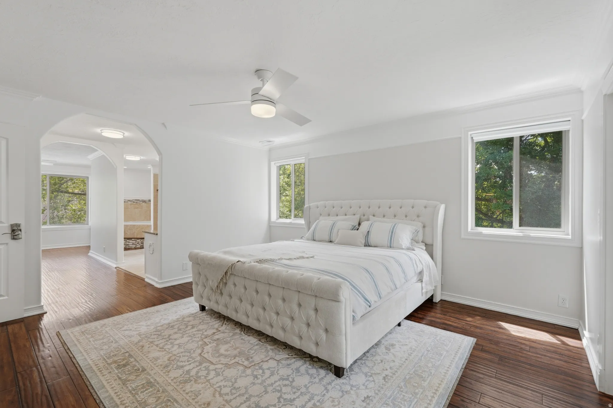 Bedroom with wood-finish flooring, two large windows, a white ceiling fan, and decorative archways leading to an additional space