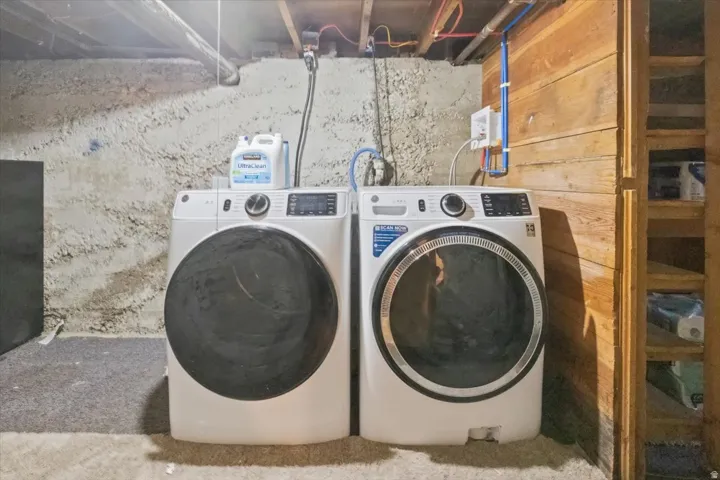 Laundry area featuring a front-loading washer and dryer, exposed wood beam ceiling, concrete wall, wood-paneled wall, and built-in wood shelving