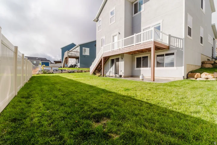 Expansive grassy yard featuring a light-colored privacy fence, a multi-level deck with white railings, a lower-level patio, and natural rock landscaping elements