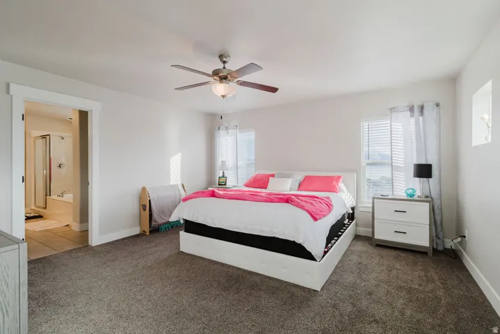 Carpeted room featuring a ceiling fan with integrated lighting, two windows with blinds, a recessed wall niche, and an adjoining tiled bathroom with a glass-enclosed shower and a built-in tub
