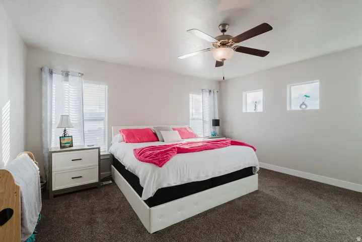 Spacious bedroom featuring neutral wall paint, dark grey carpet flooring, two windows with blinds, a ceiling fan with light fixture, and two recessed wall niches