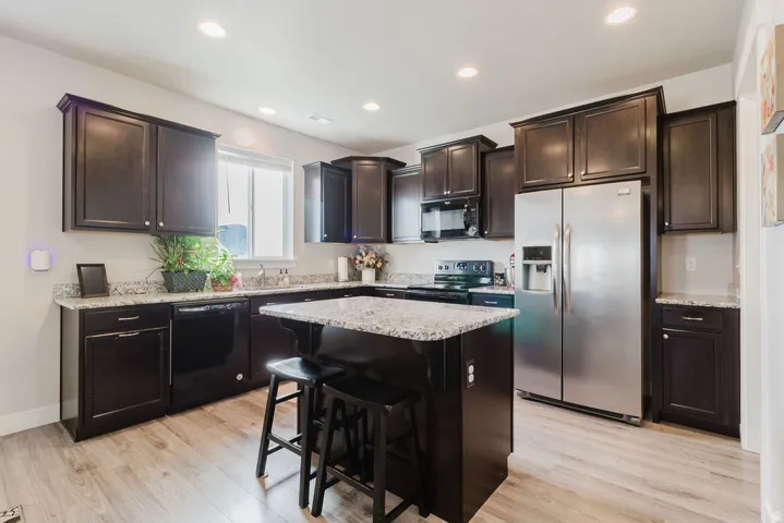 Kitchen featuring dark wood cabinetry, light-toned wood-finish flooring, a central island with a light granite countertop, stainless steel appliances, and recessed lighting