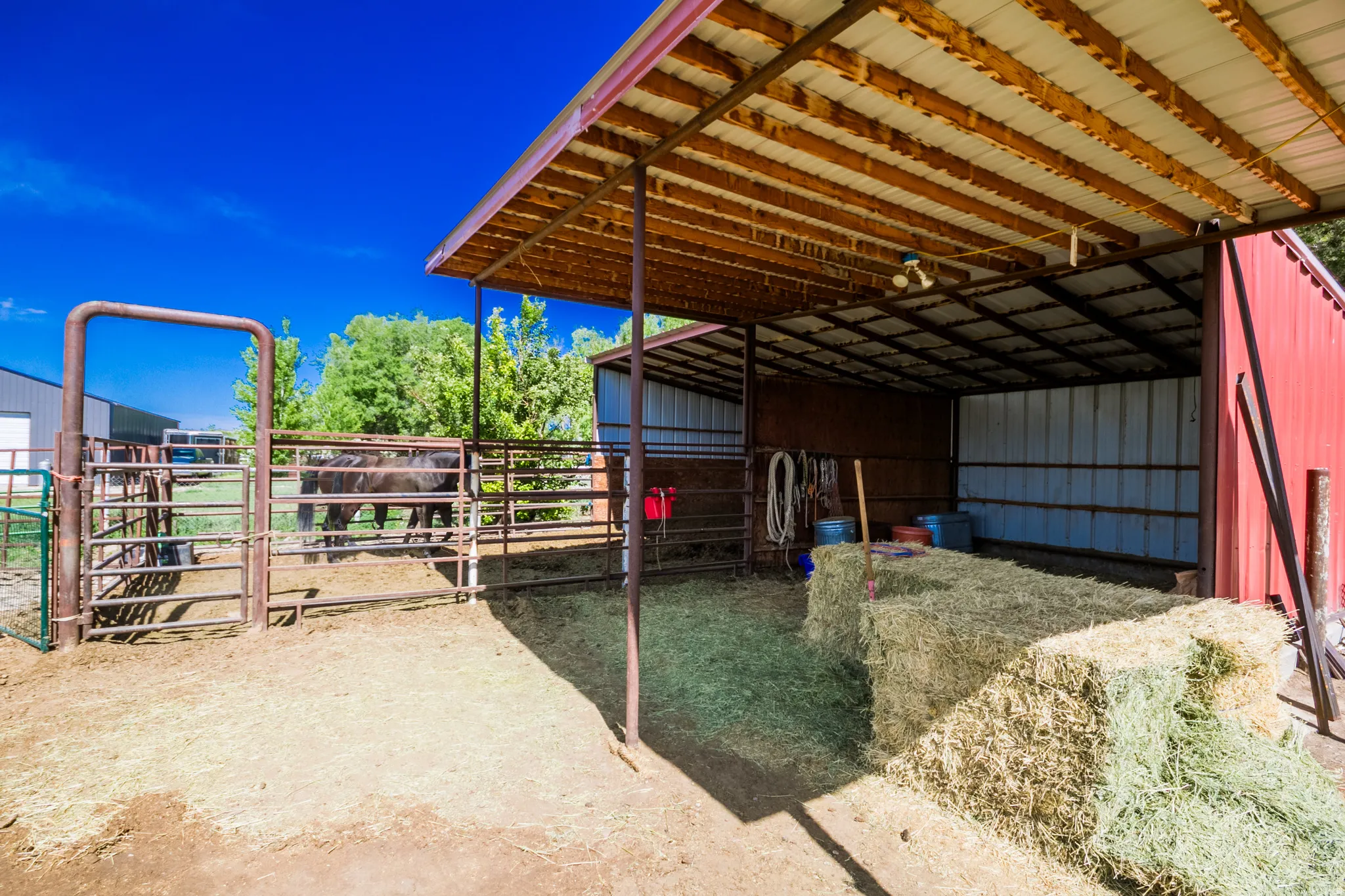 Red barn with a metal roof extension covering an open-sided stall area, featuring a dirt floor and metal fencing