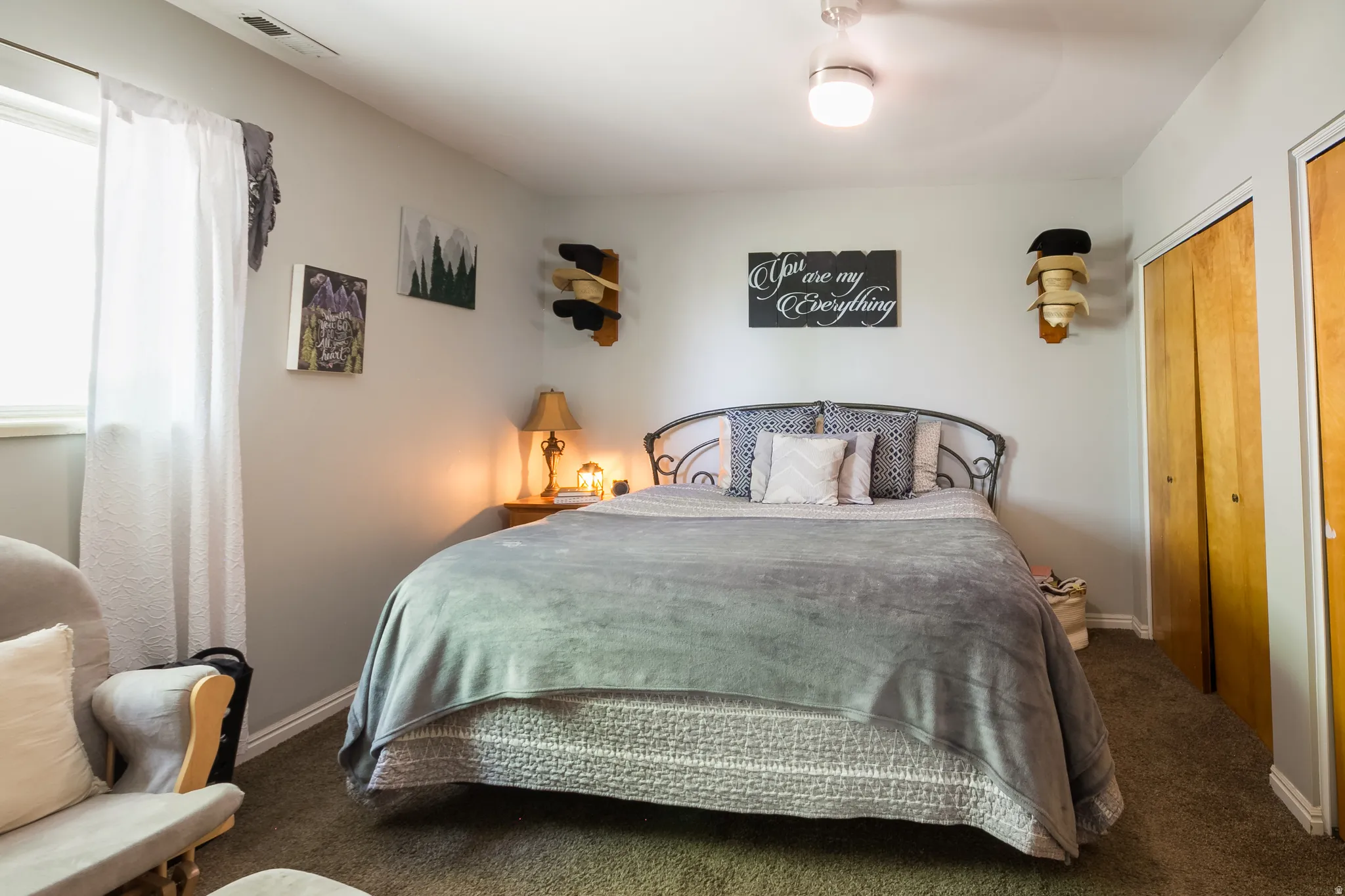 Carpeted room featuring a window with white curtains, a ceiling light fixture, a metal bed frame, and two wood-finish bi-fold closet doors
