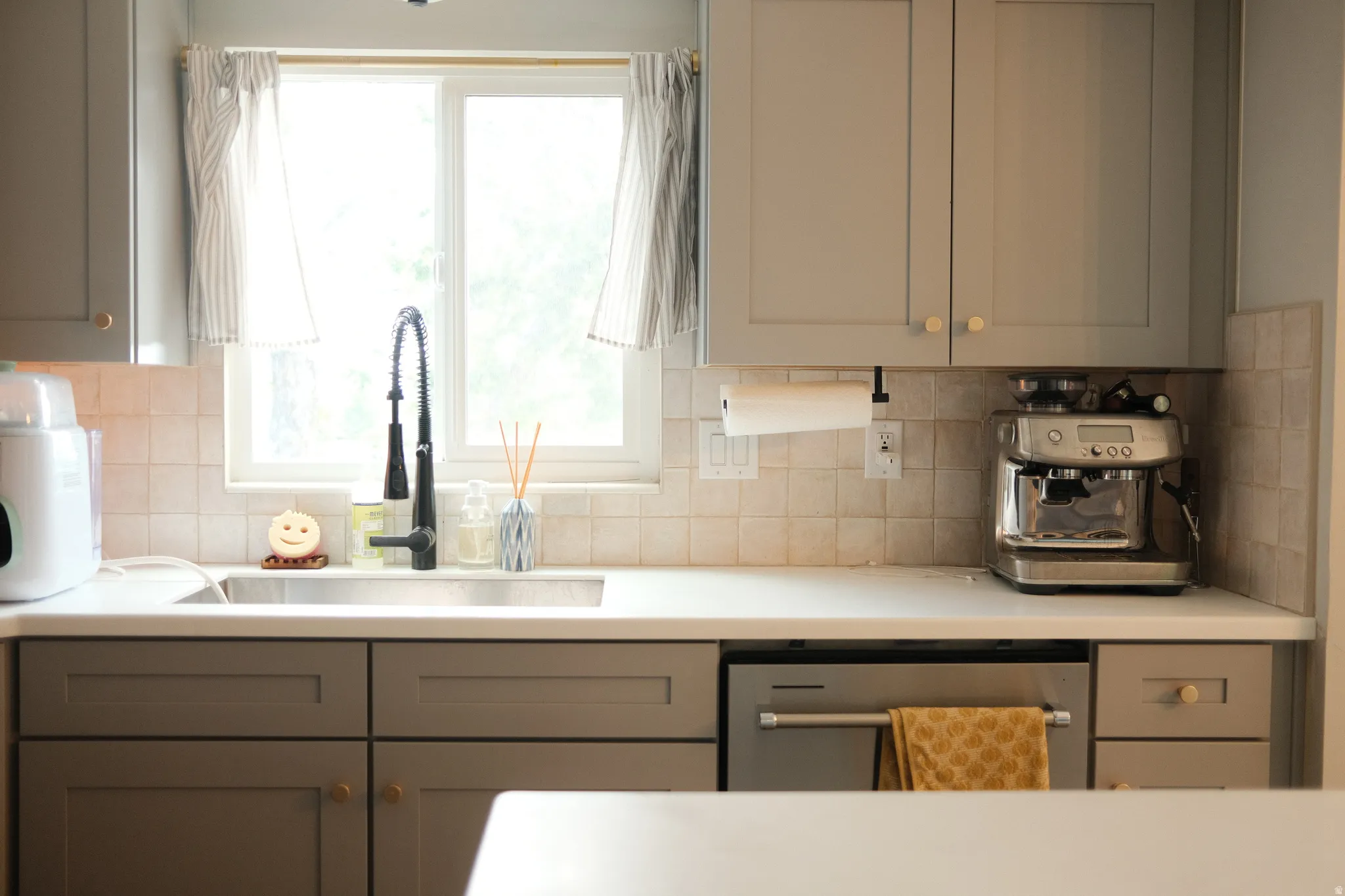 Kitchen featuring light gray shaker-style cabinetry with brass hardware, white countertops, square tile backsplash, and a stainless steel sink with a black coiled faucet