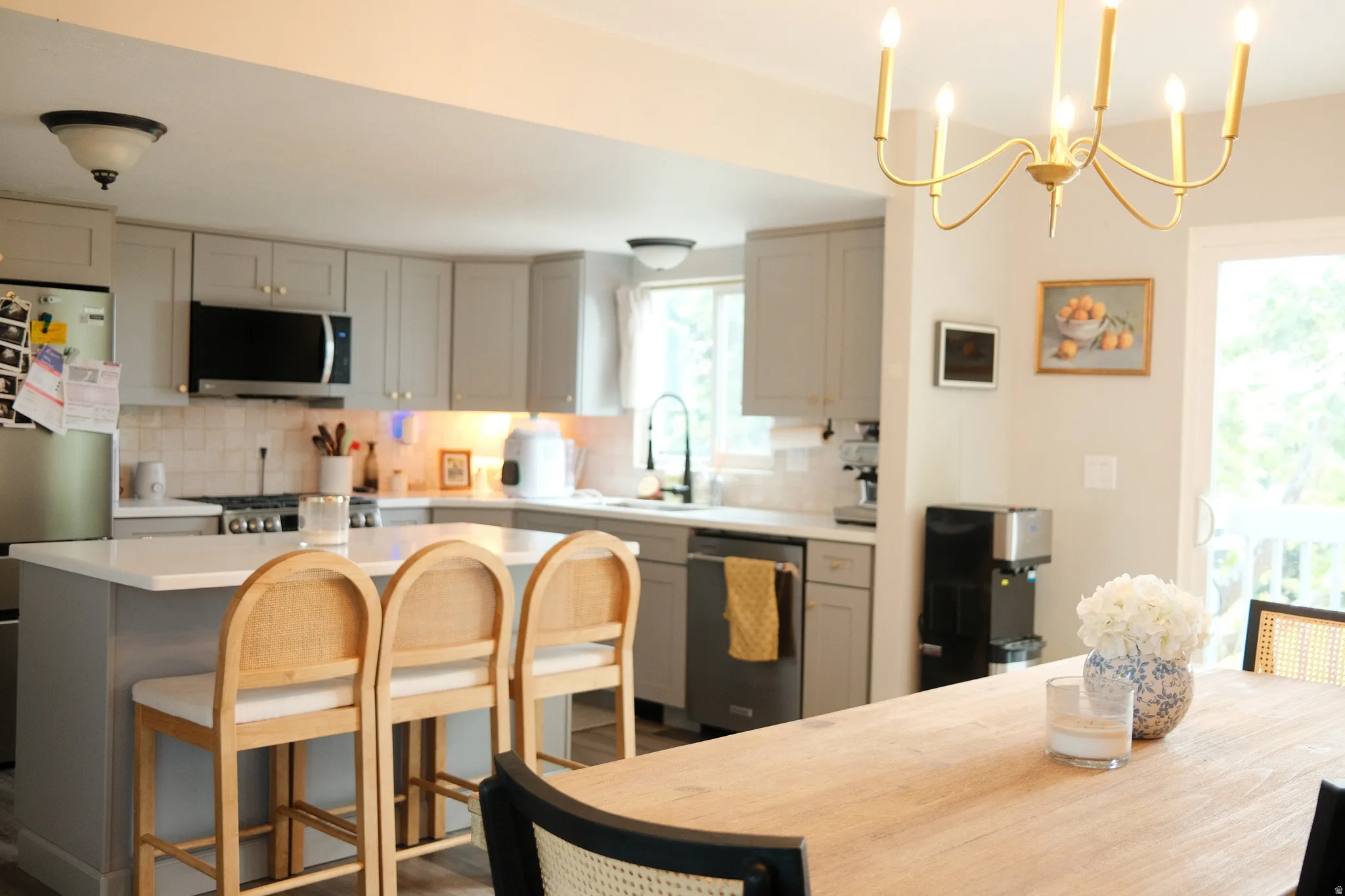 Open-concept kitchen featuring an island with built-in seating, light gray cabinetry, stainless steel appliances, a tile backsplash, and a gold-tone chandelier