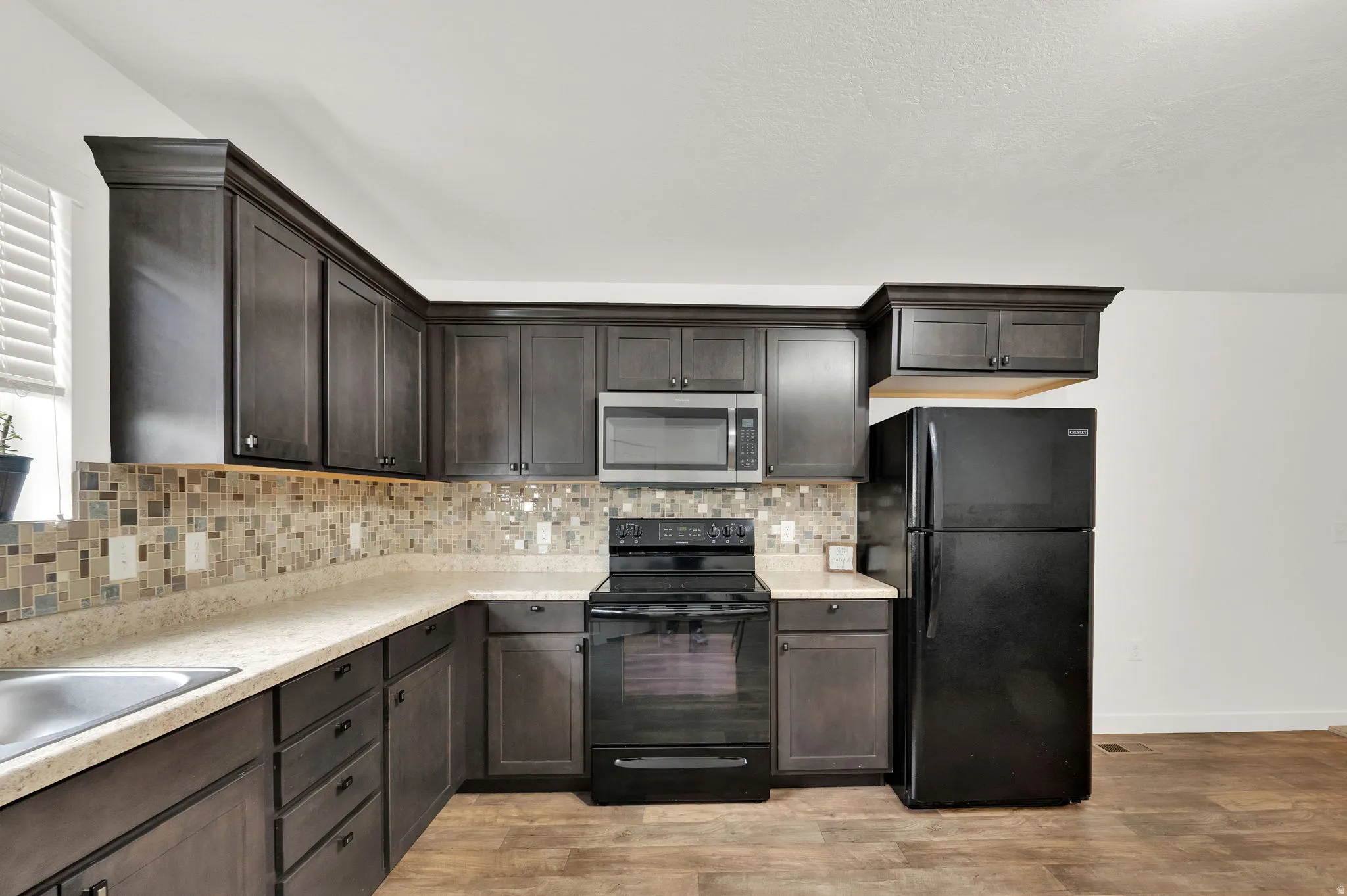 Kitchen featuring dark wood-finish cabinetry with crown molding, light-toned countertops, a mosaic tile backsplash, stainless steel microwave, and black electric range