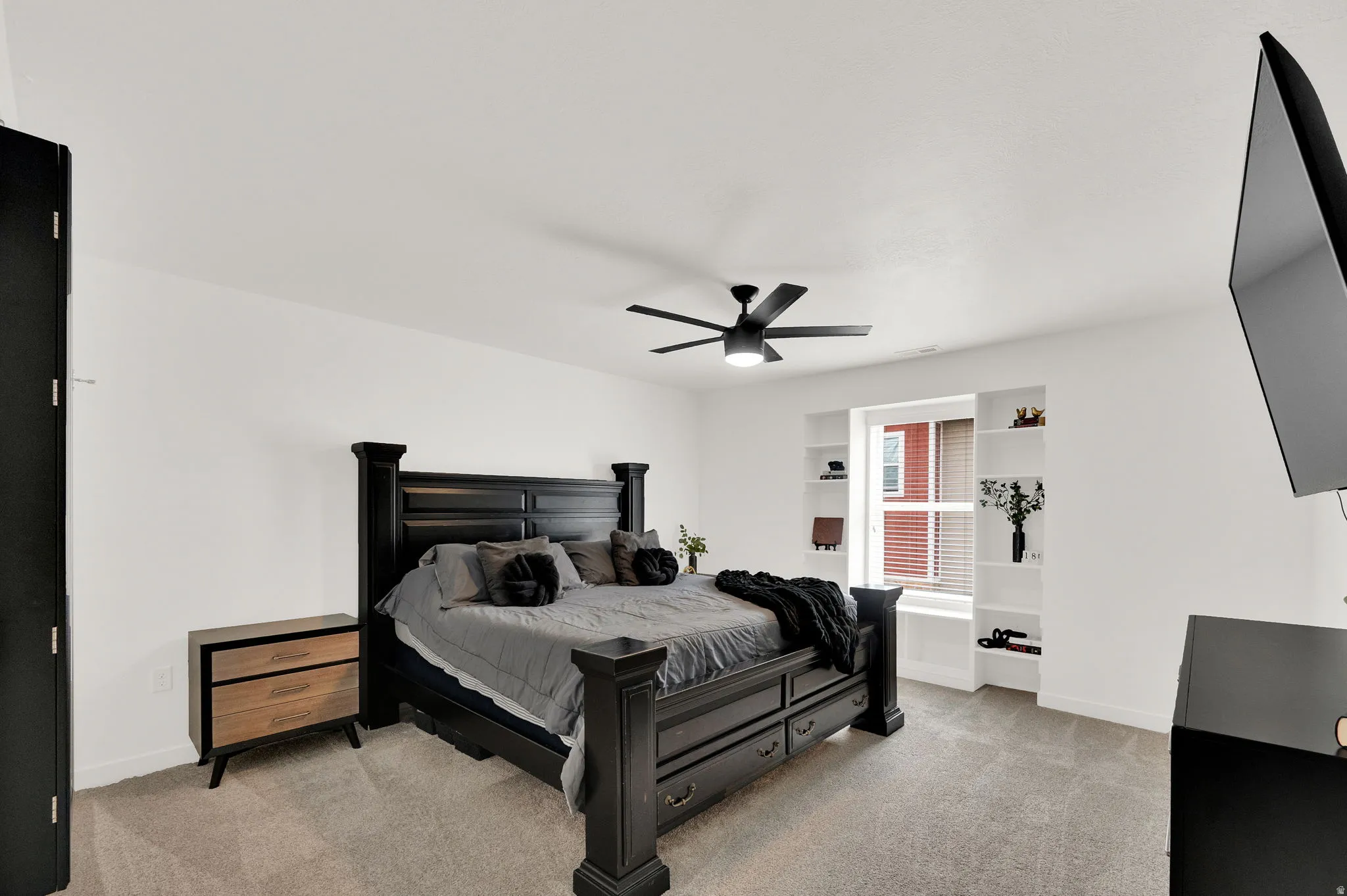 Carpeted bedroom featuring a ceiling fan with integrated lighting, a window with horizontal blinds, and built-in shelving surrounding the window