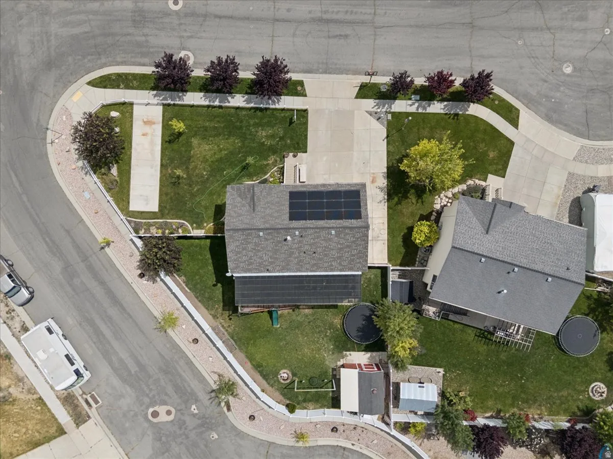 Aerial perspective of a property featuring a gray shingle roof with solar panels, a concrete driveway, and a front yard with mature trees