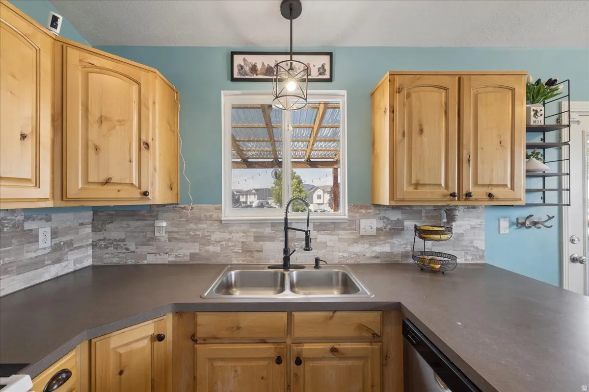 U-shaped kitchen counter featuring a double basin stainless steel sink with a matte black faucet, surrounded by wood-finish cabinetry, dark countertops, and a light-toned tile backsplash