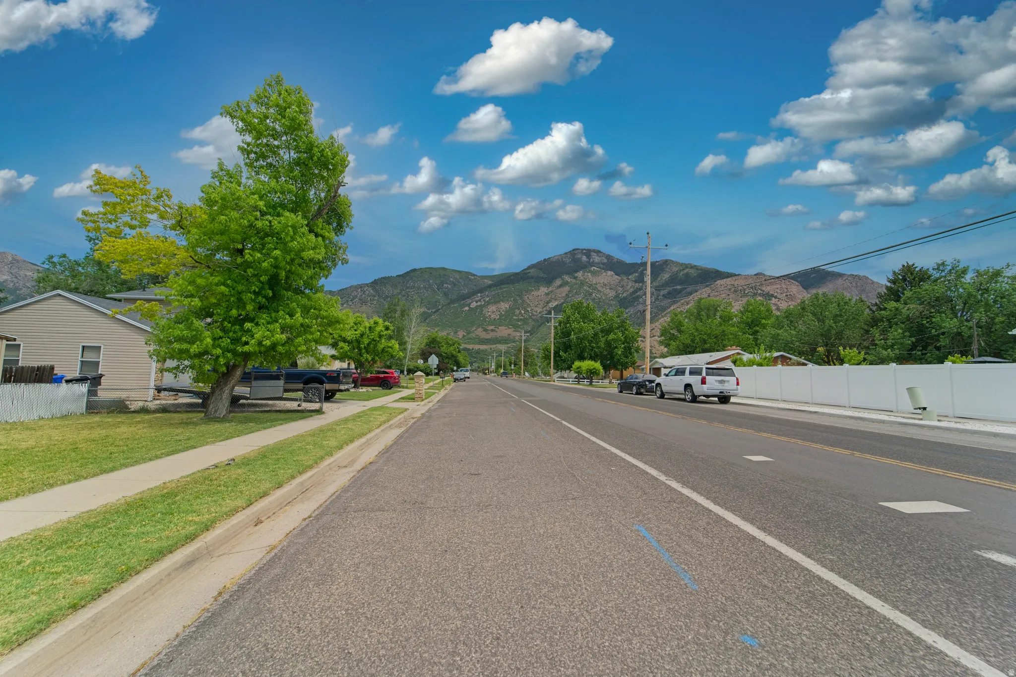 Paved street with a concrete sidewalk bordered by a grassy verge, mature trees, and a white picket fence, set against a backdrop of distant mountains