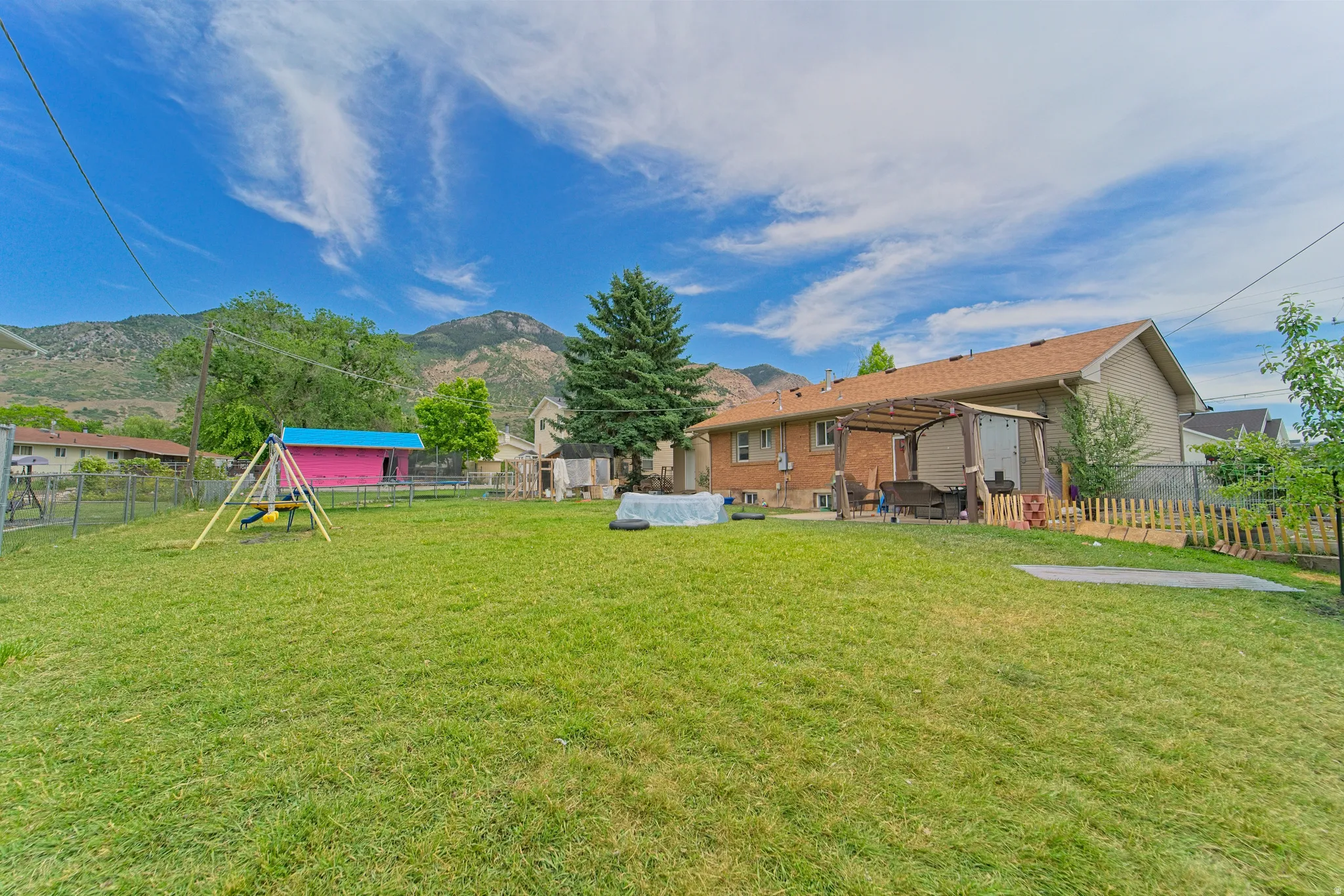 Expansive grassy backyard featuring a large patio with a wooden pergola, a chain-link fence, and a mountain backdrop