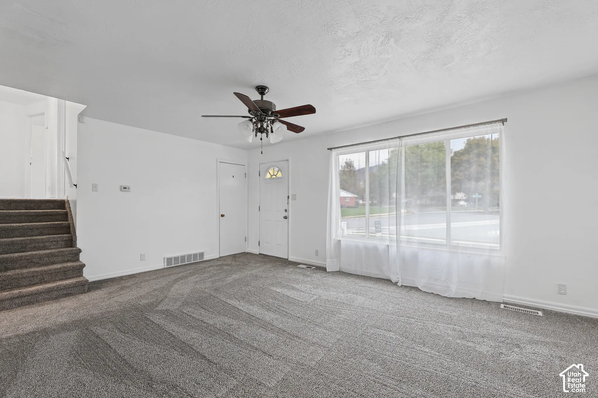 Carpeted flooring, white walls, ceiling fan, large window, and staircase with carpeted steps