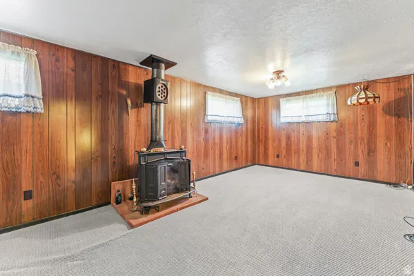 Room featuring wood-finish paneling, a classic cast iron stove with a brick-finish hearth, light-toned carpeting, and multiple windows with white curtains