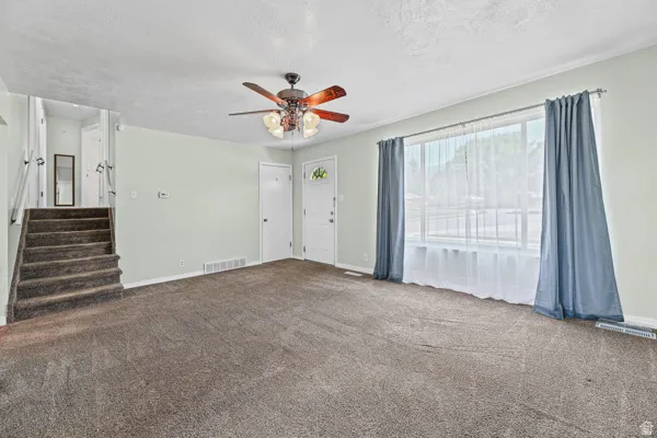 Spacious living area featuring neutral carpeting, a prominent window with privacy sheers and drapery, and a ceiling fan with integrated lighting