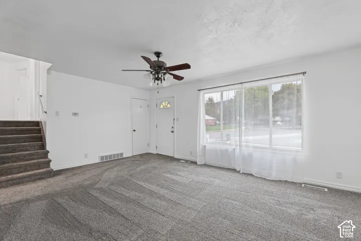 Carpeted flooring, white walls, ceiling fan, large window, and staircase with carpeted steps