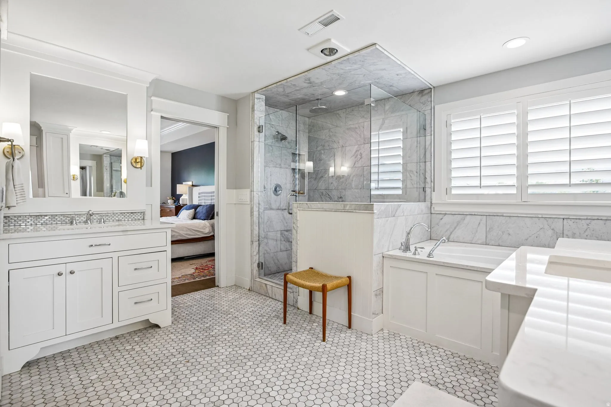 Bathroom featuring white hexagon tile flooring, a glass-enclosed shower with tiled walls, a separate soaking tub with a tiled surround, white vanity cabinetry with a stone countertop, and plantation shutters