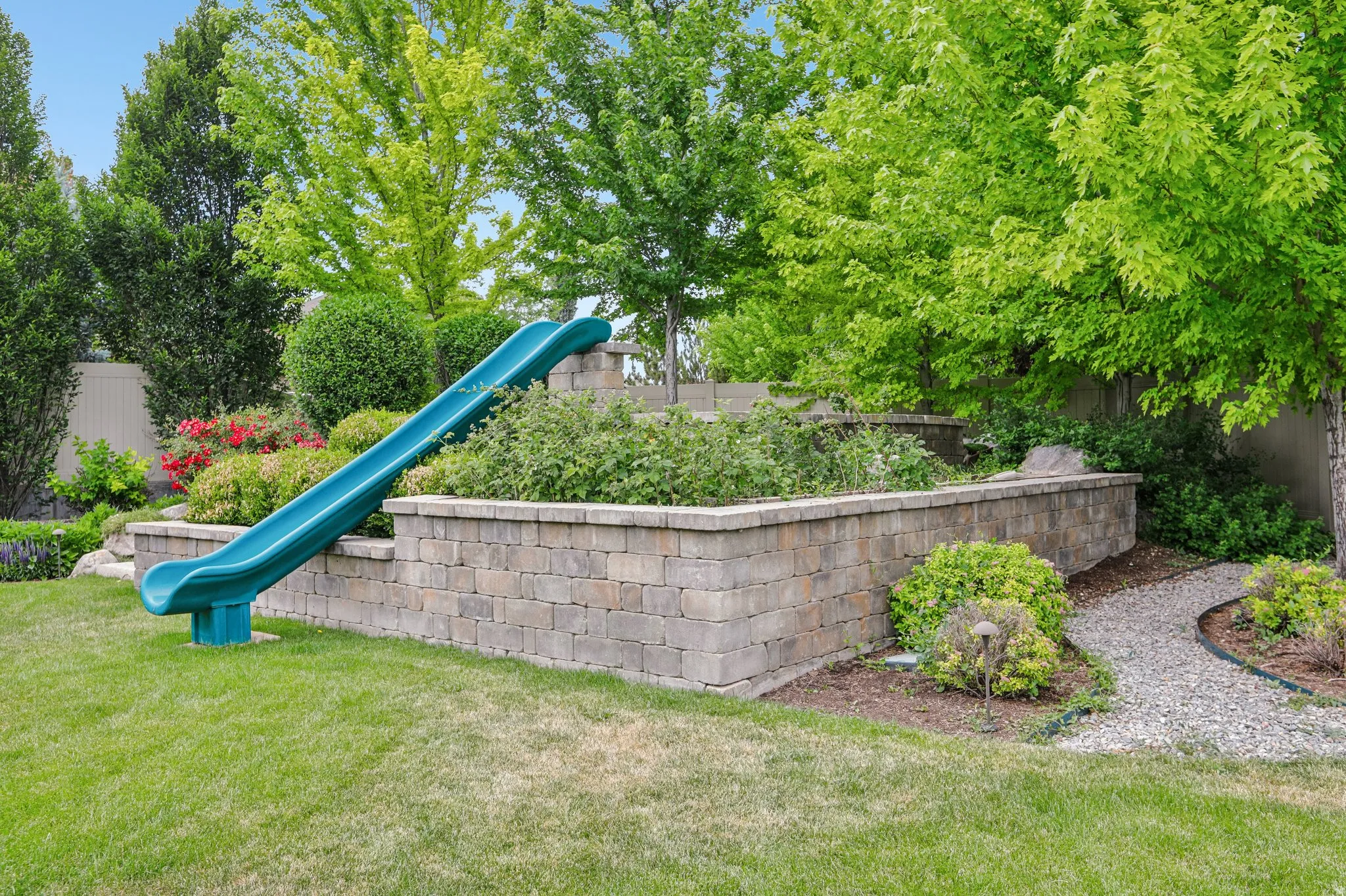 Expansive backyard featuring tiered retaining walls constructed from paver blocks, a gravel pathway, and a variety of mature trees and shrubs