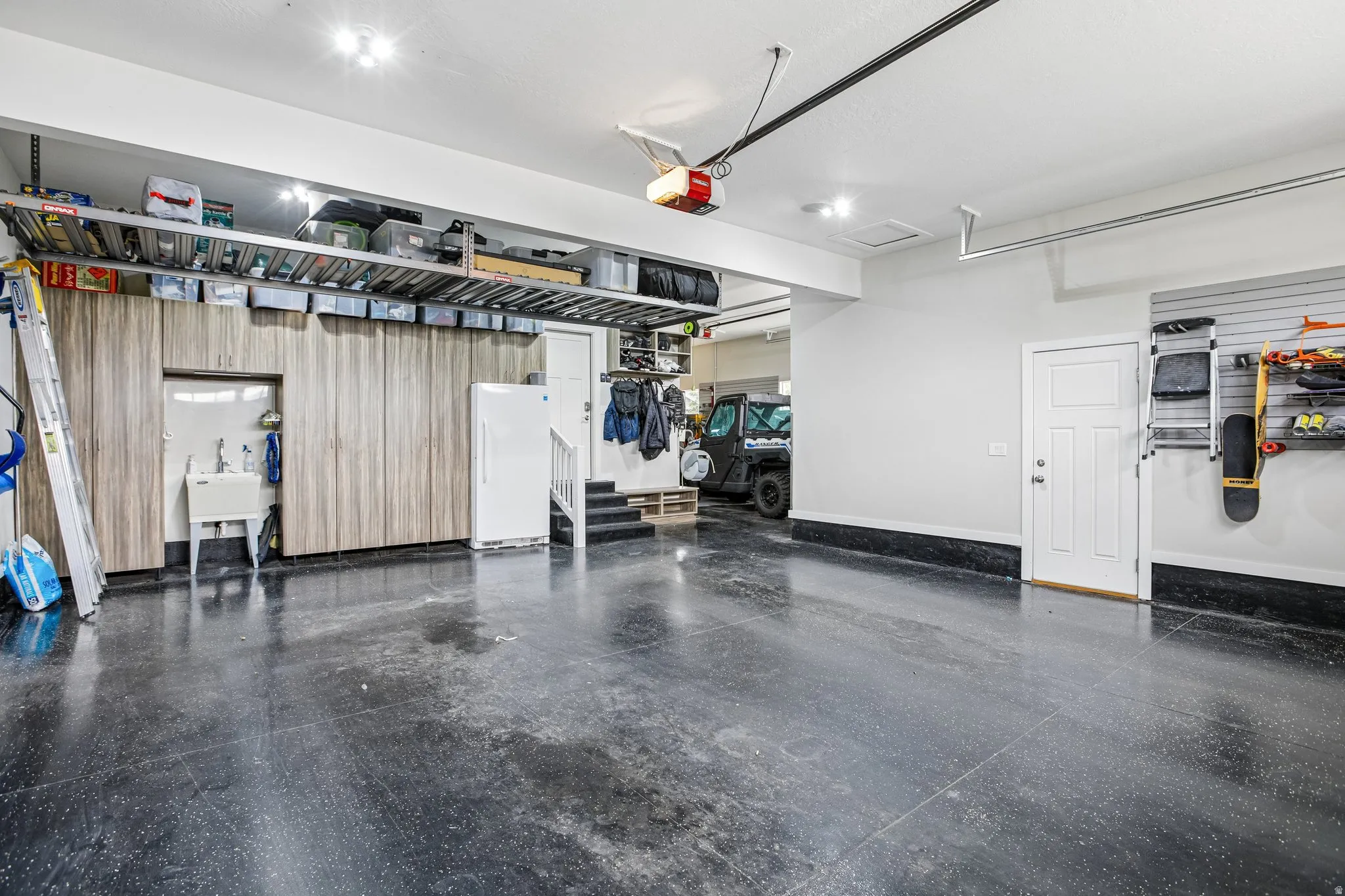 Spacious garage featuring a durable dark flooring finish, extensive built-in wood-finish cabinetry, an integrated utility sink, overhead storage racks, and a white entry door