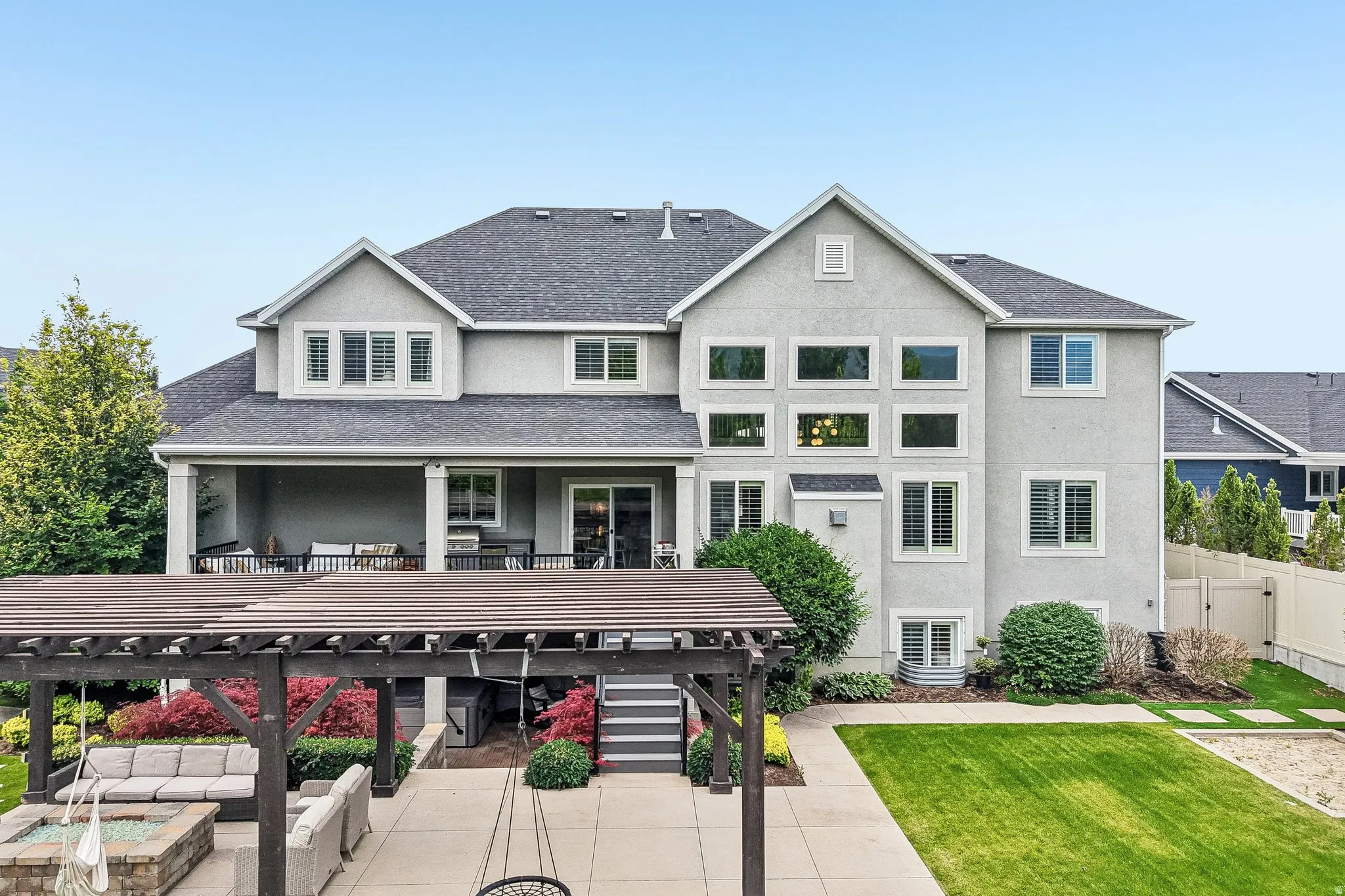 Rear exterior featuring a large stucco facade, multiple window configurations, a covered patio with white columns, a dark wood pergola over a paved patio, and a manicured lawn