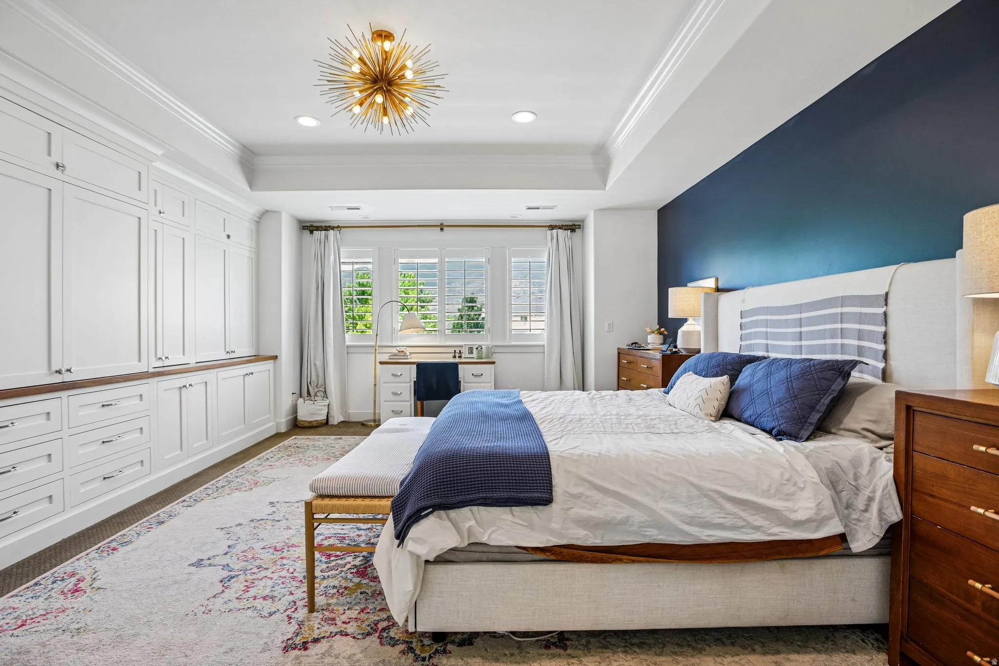 Recessed ceiling with a modern light fixture, extensive white built-in cabinetry with wood-finish countertops, custom window shutters, and a contrasting accent wall