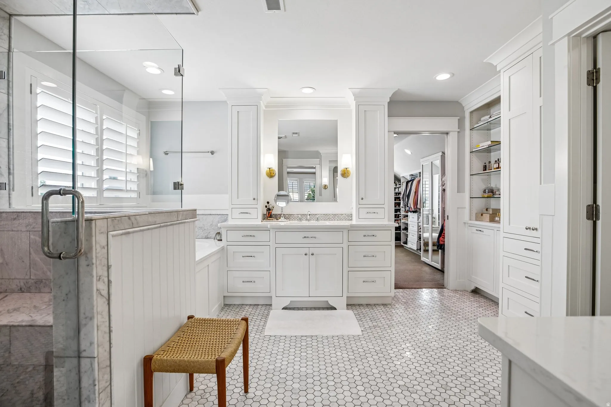 Spacious bathroom featuring white hexagonal tile flooring, a glass-enclosed shower, a built-in soaking tub, a double vanity with stone countertops, and extensive built-in cabinetry with open shelving