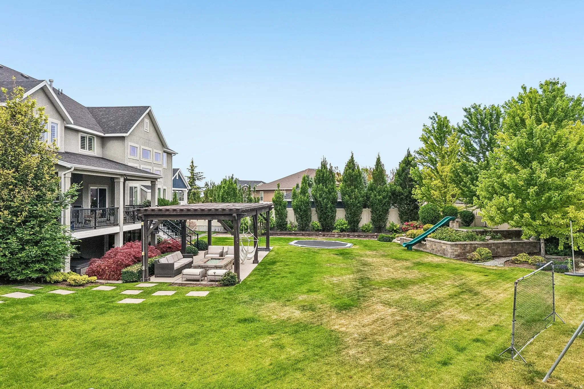 Expansive backyard featuring a large lawn area, a paved patio with a wooden pergola, a raised garden bed with a slide, and mature trees providing natural screening