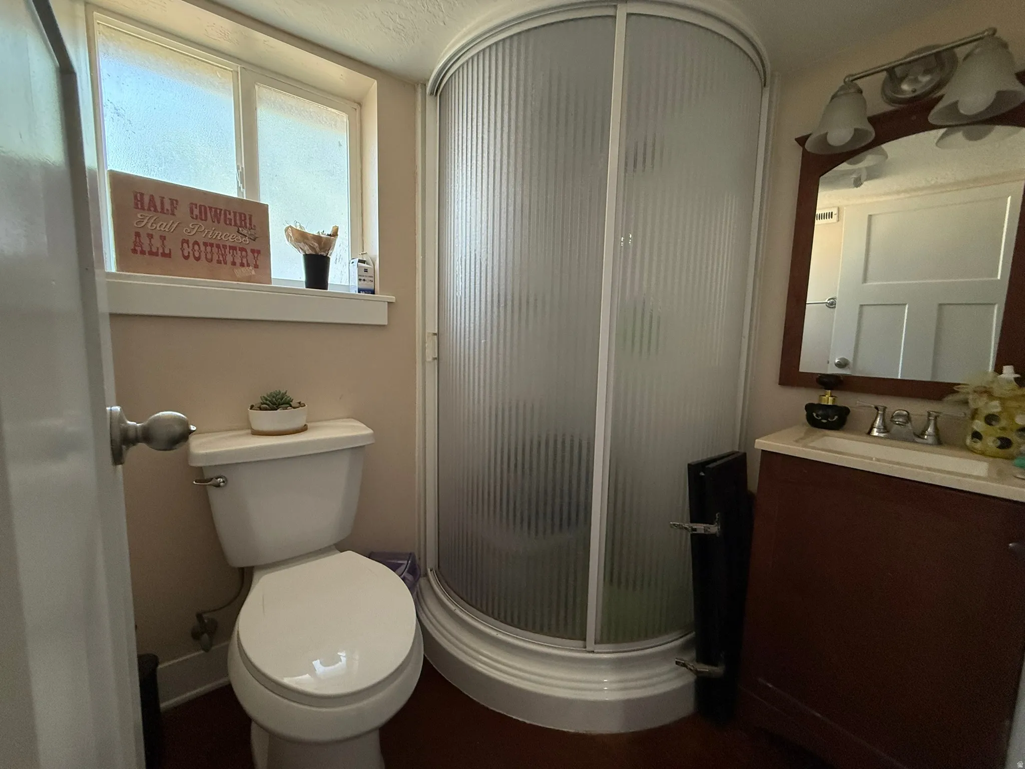 Compact bathroom featuring a curved shower enclosure, single vanity with a cream-toned countertop, wood-finish flooring, and a window with frosted glass