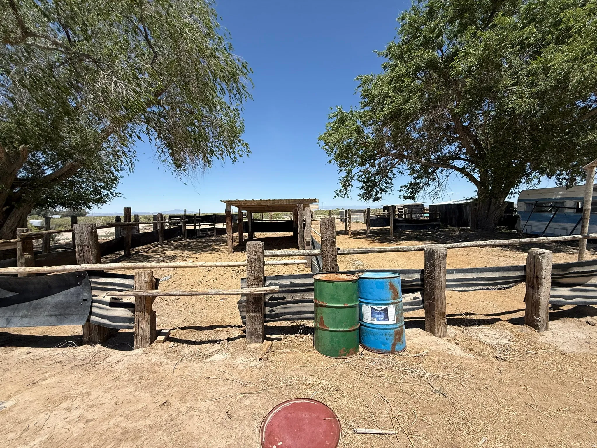 Fenced outdoor area featuring a wooden post and rail fence, a corrugated metal barrier, and mature trees providing natural shade