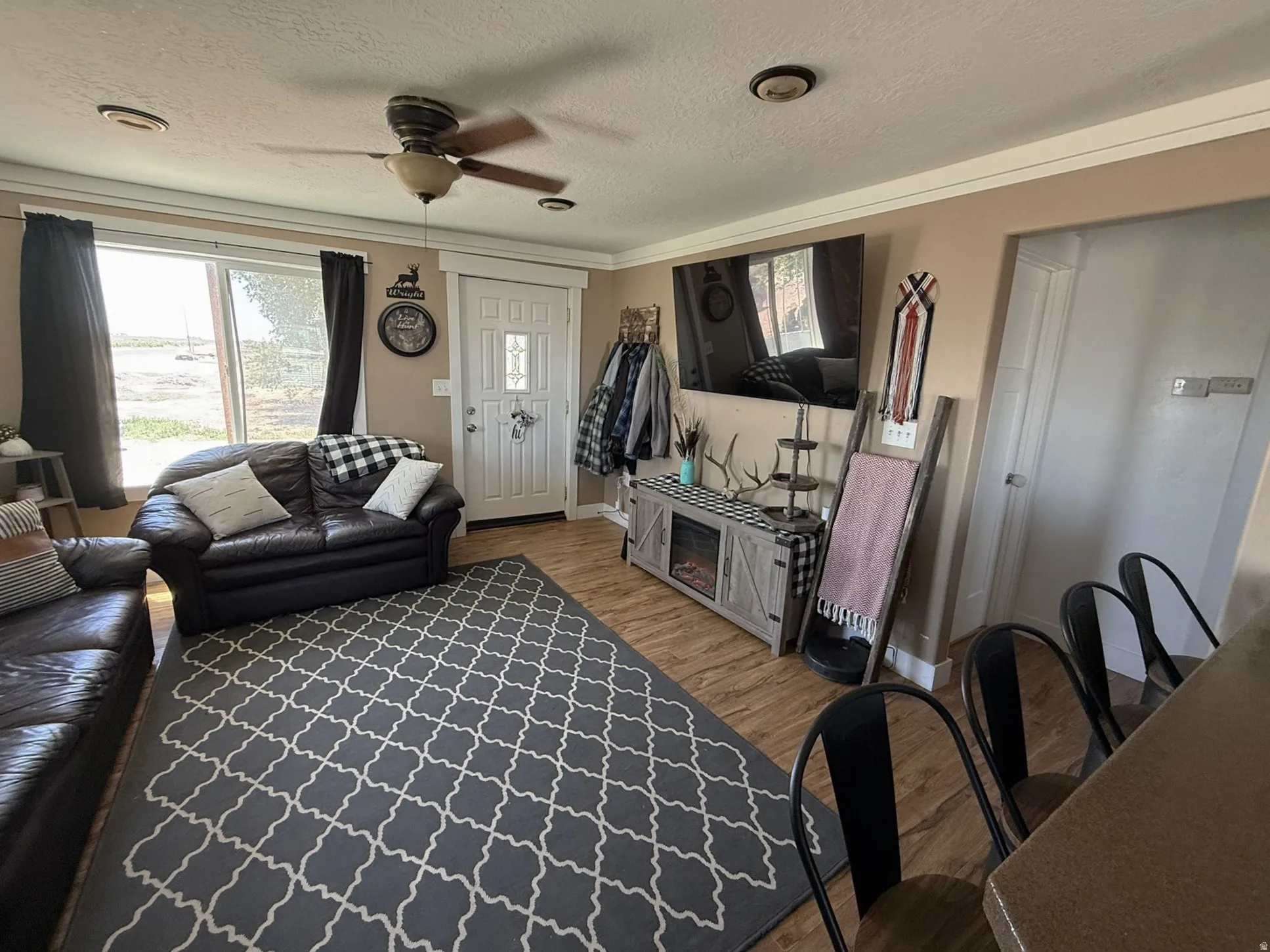 Light-filled living area featuring wood-finish flooring, a ceiling fan, crown molding, and a built-in entertainment console with a simulated fireplace