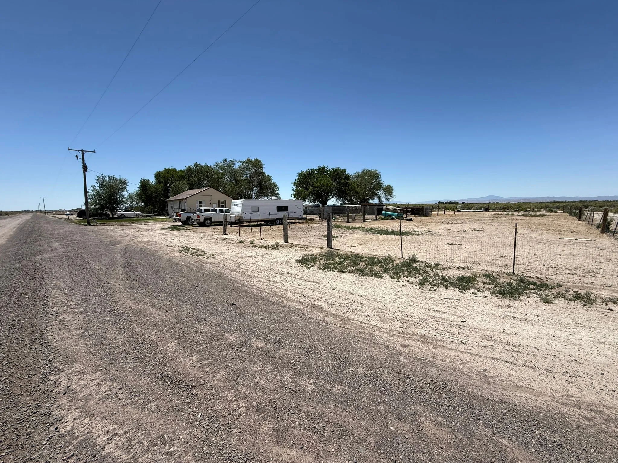Expansive gravel lot featuring a single-story residence with light-toned siding, mature trees providing natural shading, and perimeter fencing