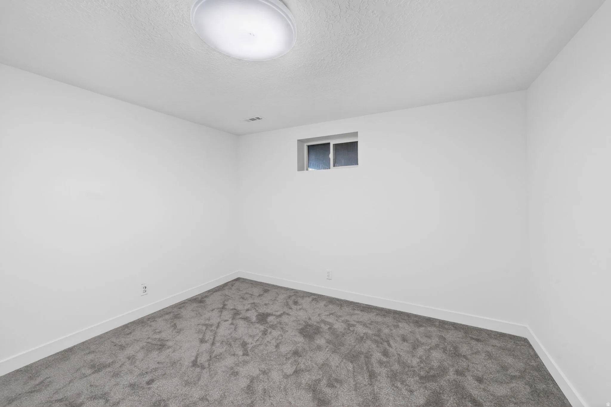 Spacious room featuring neutral white walls, plush gray carpeting, white trim, and a high-mounted window