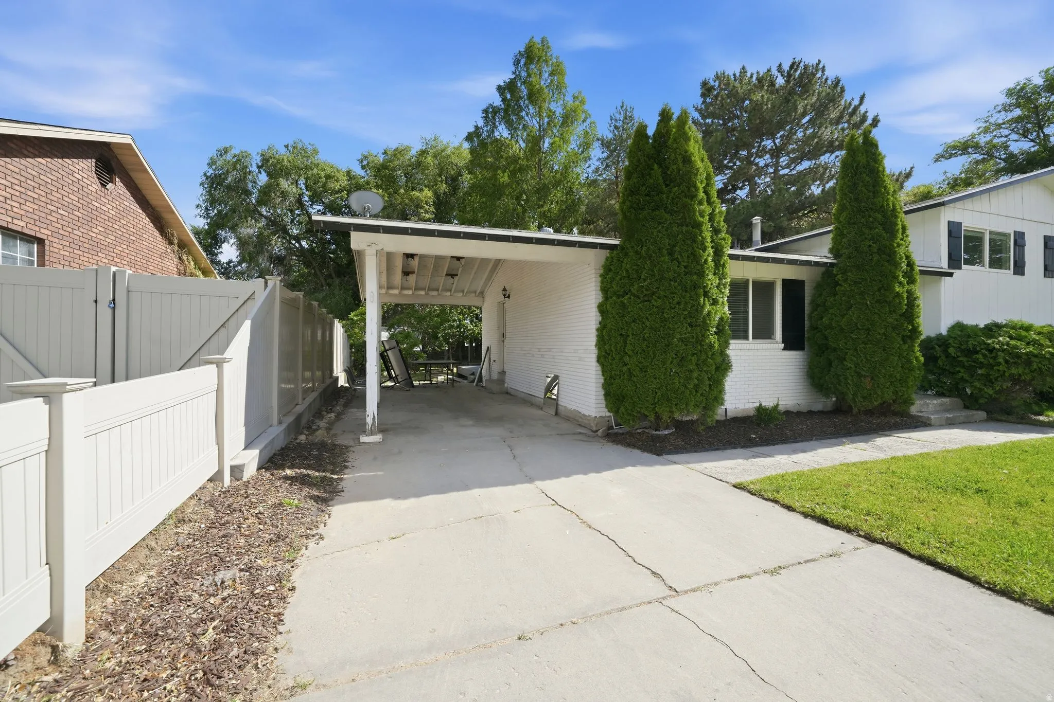 White painted brick exterior with a covered carport, concrete driveway, manicured lawn, and mature evergreen shrubs