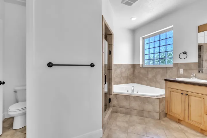 Bathroom featuring a corner soaking tub, a block glass window, tile flooring, a wood-finish vanity with a built-in sink, and a separate toilet area