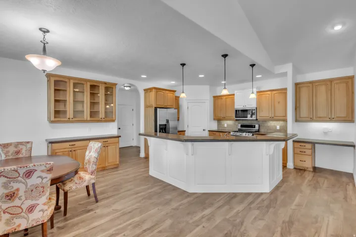 Open-concept kitchen and dining area featuring a central island with pendant lighting, wood-finish flooring, light wood cabinetry with glass-front accents, and built-in desk space