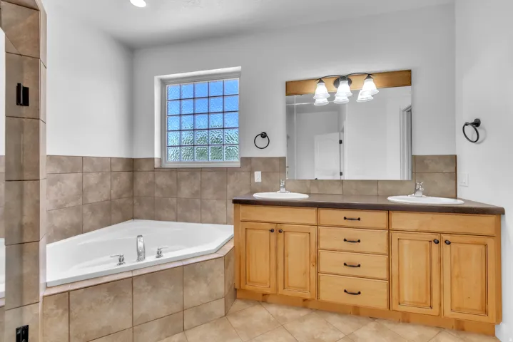 Spacious bathroom featuring a corner soaking tub with tile surround, a dual vanity with light-toned cabinetry and a dark countertop, and a glass block window providing natural light