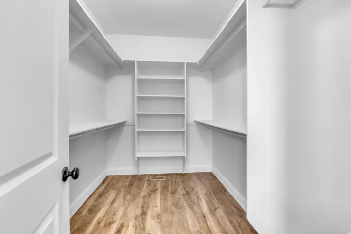 Walk-in closet featuring wood-finish flooring, white built-in shelving, white walls, and a white six-panel door with a black knob