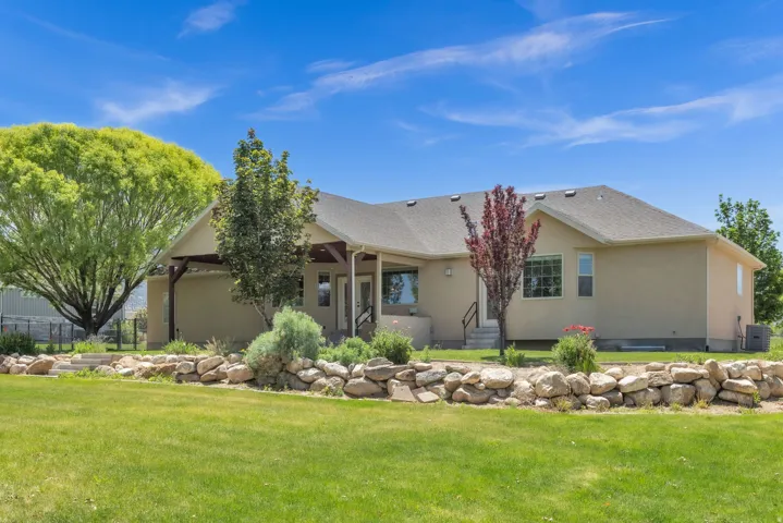 Rear exterior featuring a stucco facade, covered patio with wood beams, rock retaining wall, and mature landscaping
