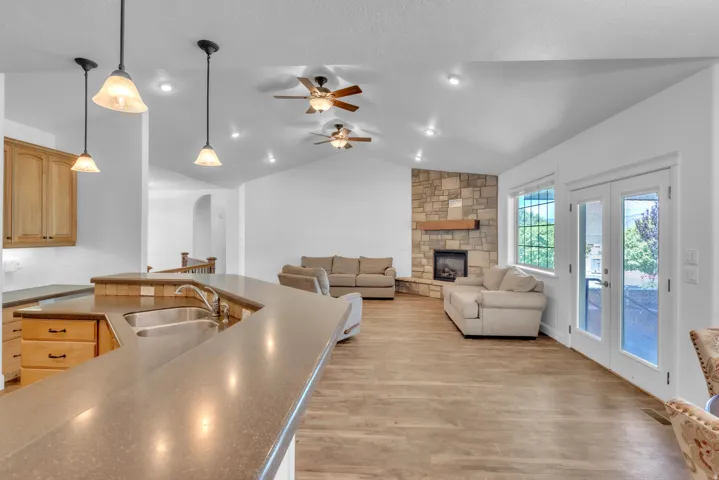 Vaulted ceilings enhance this open-concept space featuring wood-finish flooring, a stone-faced fireplace with a wood mantel, and large double-pane French doors