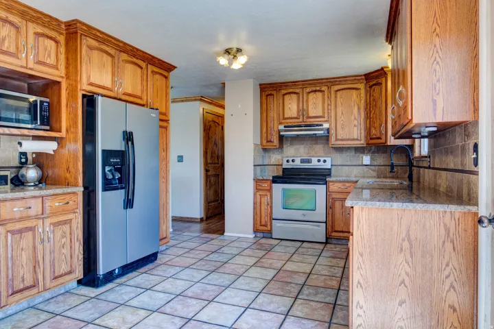Kitchen featuring wood cabinetry, stainless steel appliances, tile flooring, stone-finish countertops, and a tile backsplash