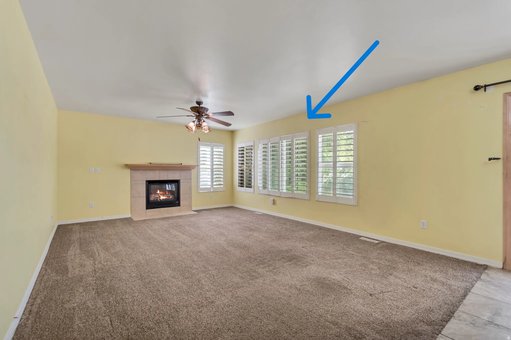 Spacious room featuring carpeting, an electric fireplace with and mantle, multiple windows with white plantation shutters (blue arrow), and a ceiling fan