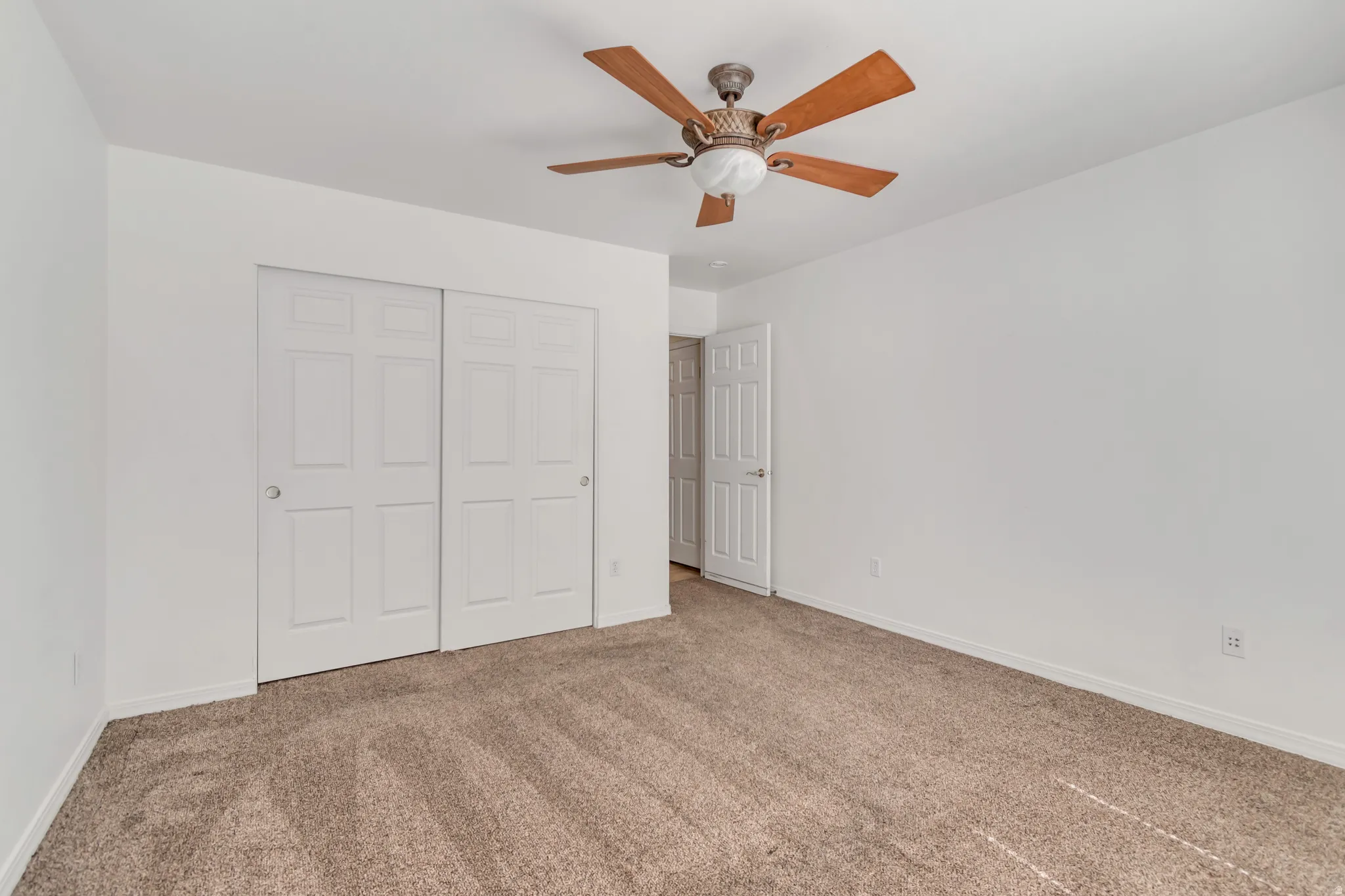 Spacious room featuring neutral carpeting, white walls, a ceiling fan with wood-finish blades, and a double sliding closet with panel doors