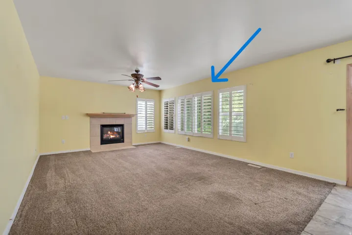 Spacious room featuring carpeting, an electric fireplace with and mantle, multiple windows with white plantation shutters (blue arrow), and a ceiling fan