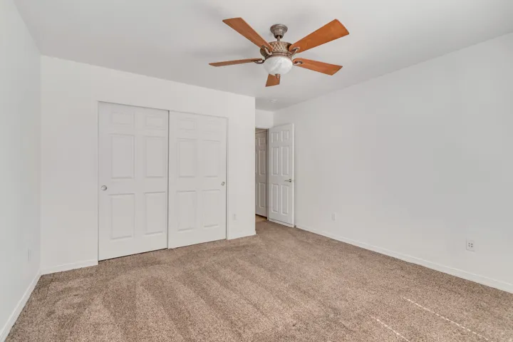 Spacious room featuring neutral carpeting, white walls, a ceiling fan with wood-finish blades, and a double sliding closet with panel doors