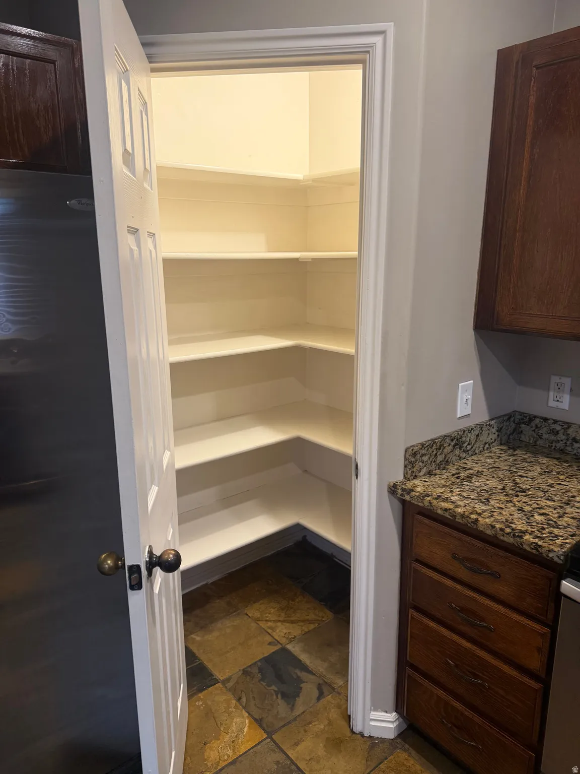 Walk-in pantry with multiple corner shelves, slate tile flooring, and a paneled door with bronze hardware