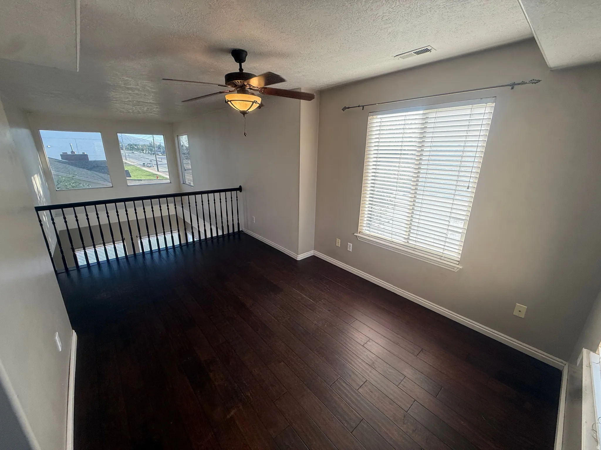 Dark wood-finish flooring, ceiling fan with integrated lighting, multiple windows with blinds, and wrought iron railing